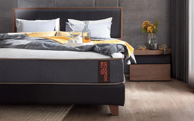 Modern bedroom with a neatly made bed featuring an orthopedic mattress, grey bedding with yellow accents, and a wooden nightstand with flowers